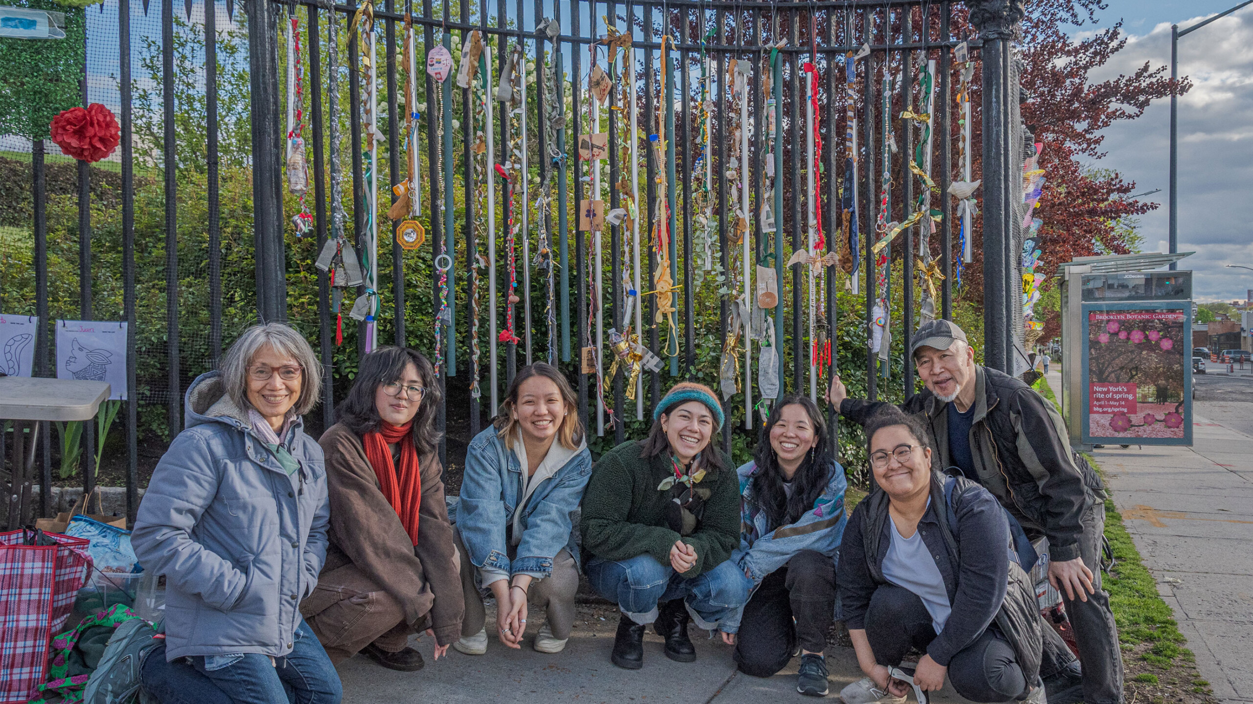 Seven people are gathered closely together, posing for a group photo while squatting or kneeling on a pavement. They are in front of a black metal fence adorned...