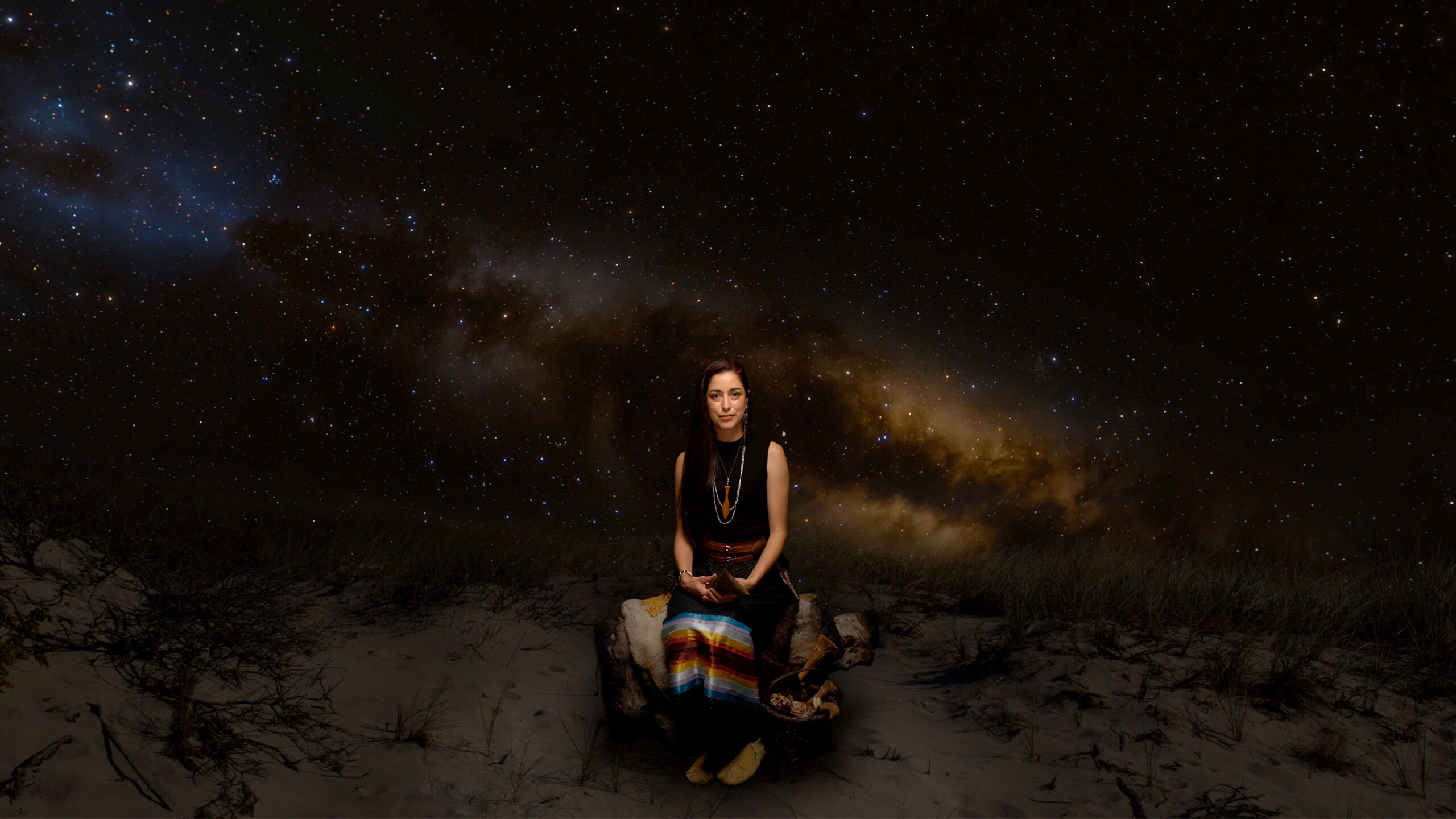 A woman sits serenely on rocky terrain under a vast, starry sky, highlighted by the brilliant Milky Way stretching across the night. Her posture is calm and meditative,...