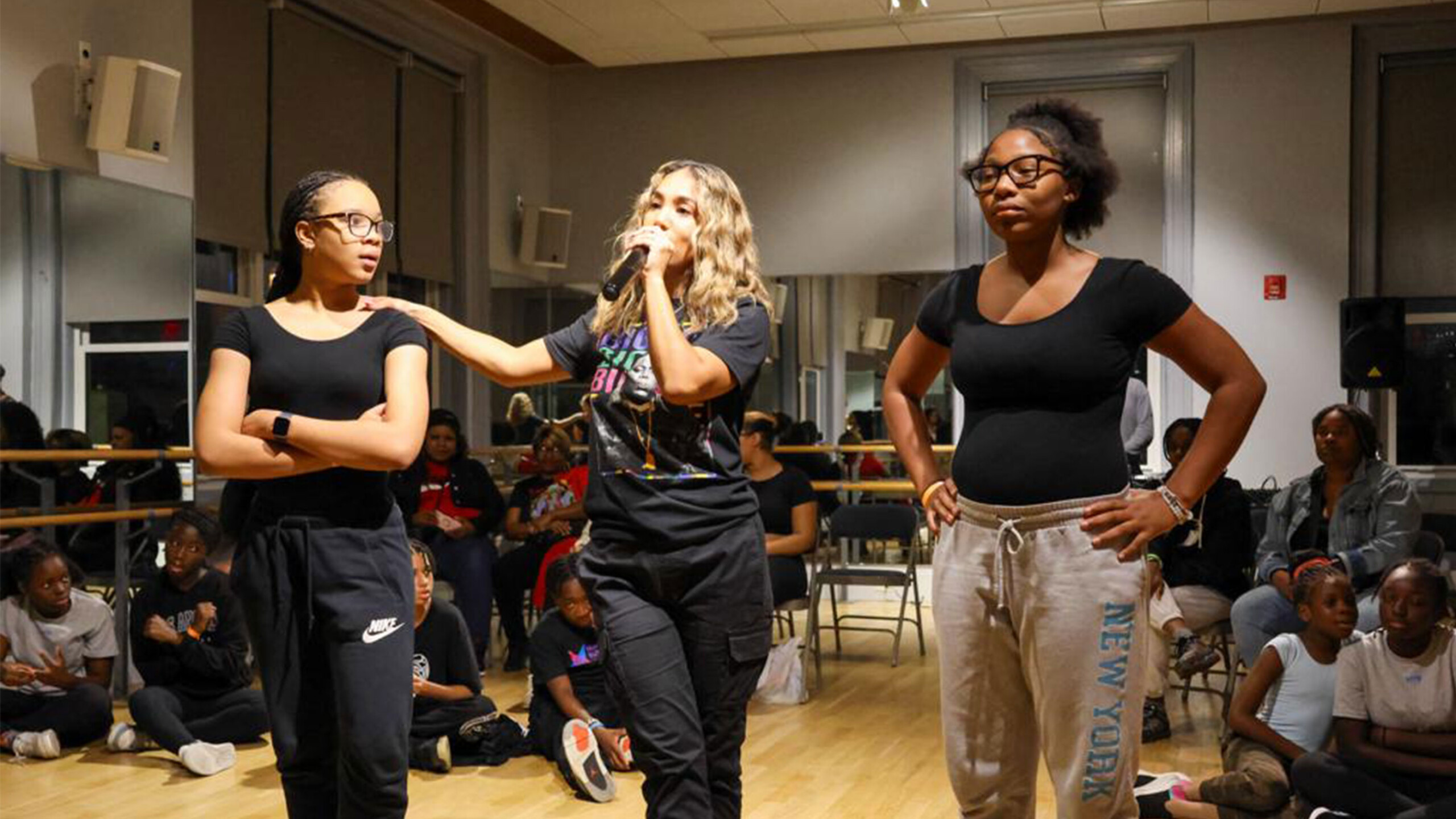Three individuals stand confidently in a dance studio with an audience in the background.