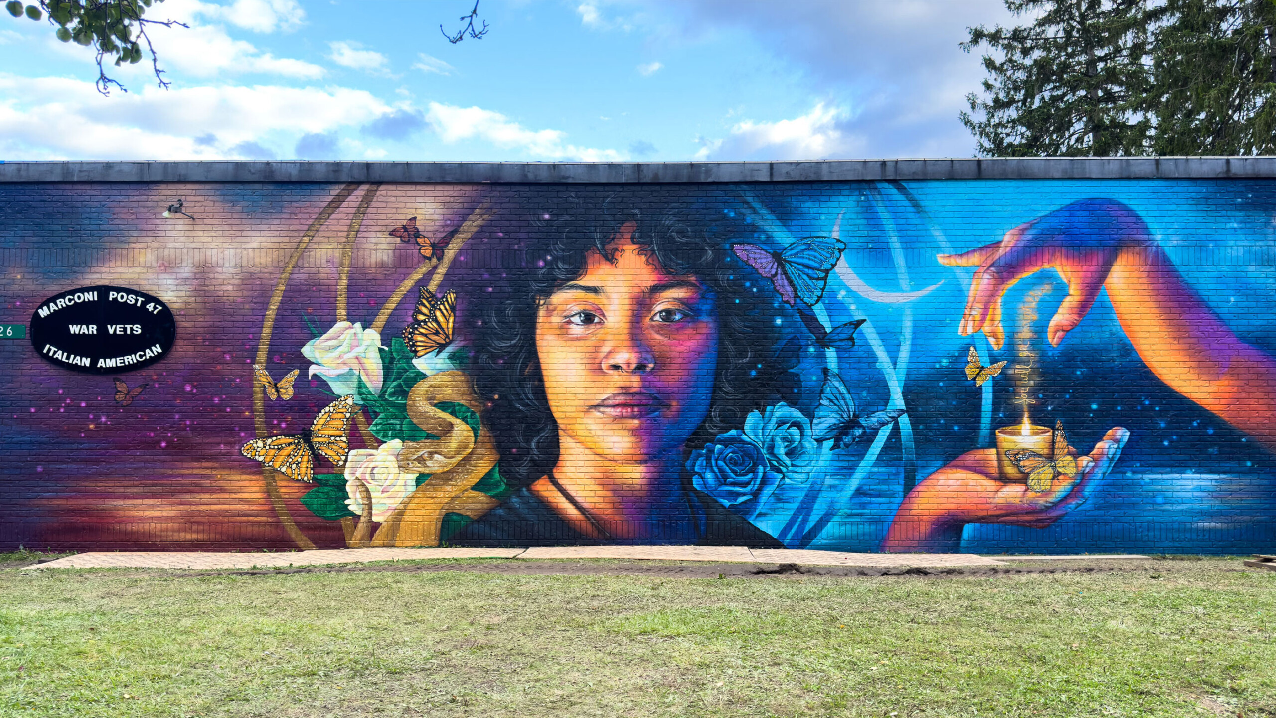 Full outdoor shot of a large mural painted on a brick wall. The mural prominently features a portrait of a woman with dark, curly hair. Her face is turned slightly...