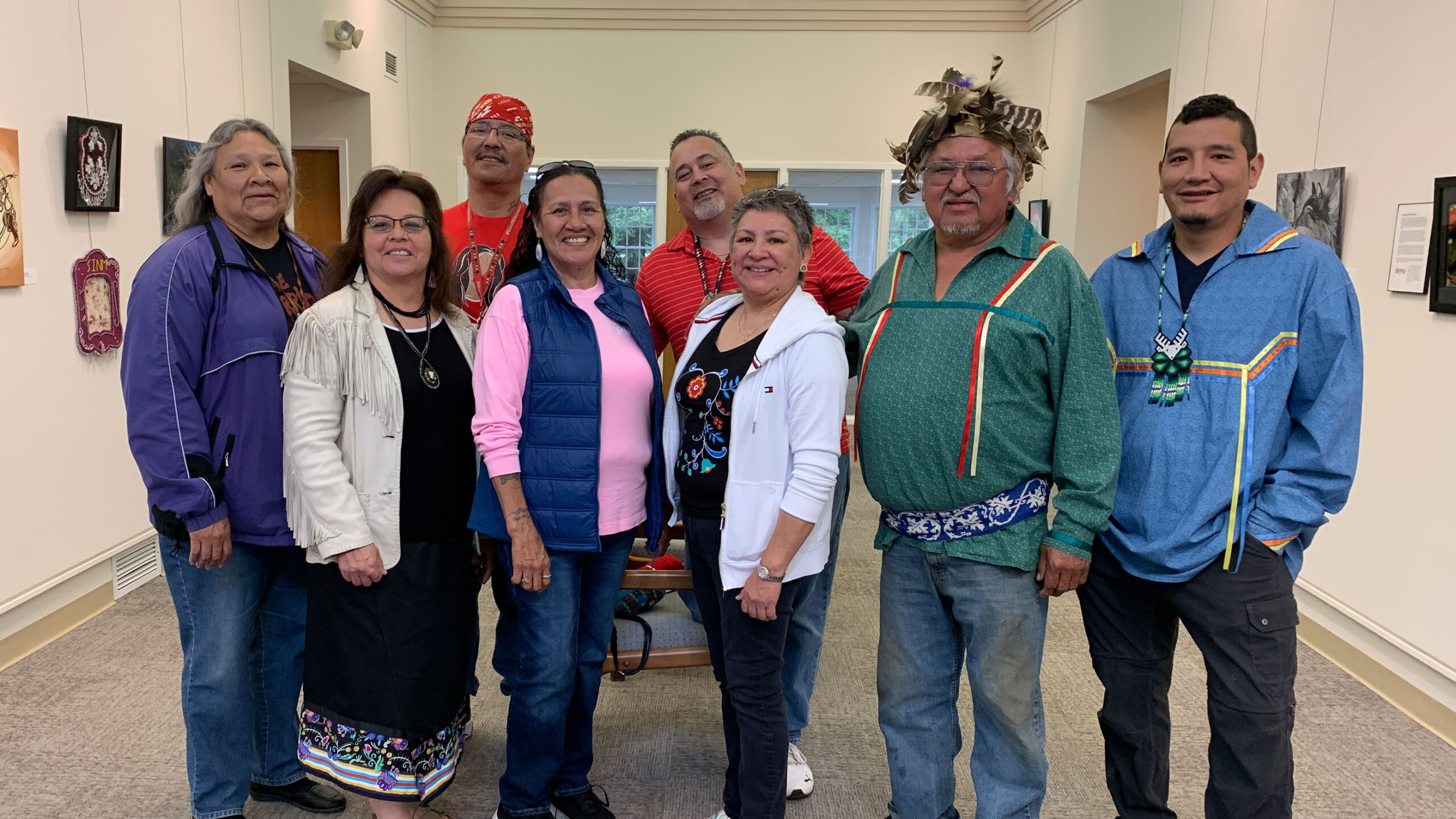 A group of eight adults stand in a well-lit room, arranged in two rows. They are wearing a mix of traditional and casual clothing, with vibrant colors and patterns....