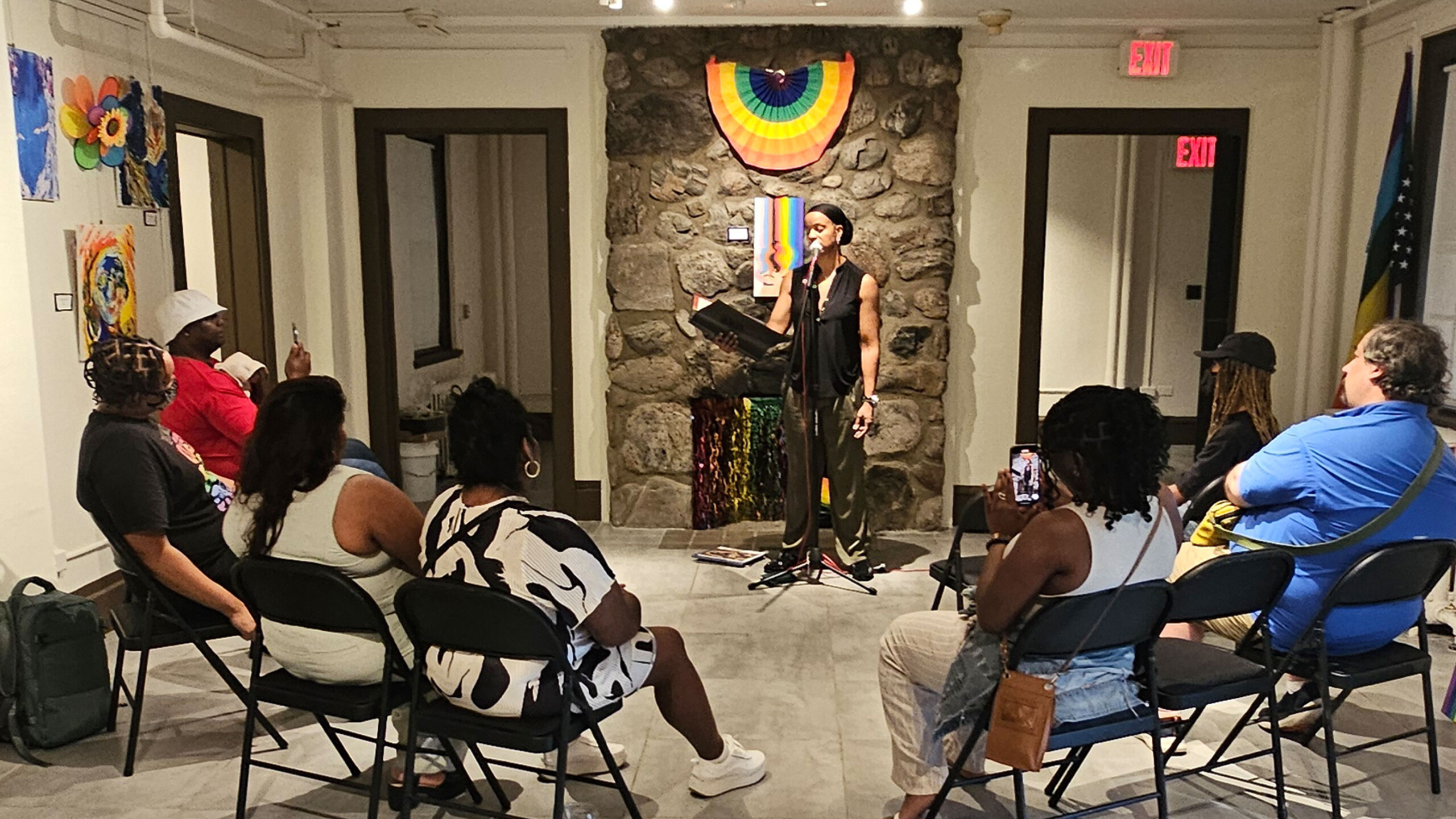A person stands in front of a stone wall, holding a book and reading aloud in a small, intimate setting. The audience, seated in a semi-circle on foldable chairs,...