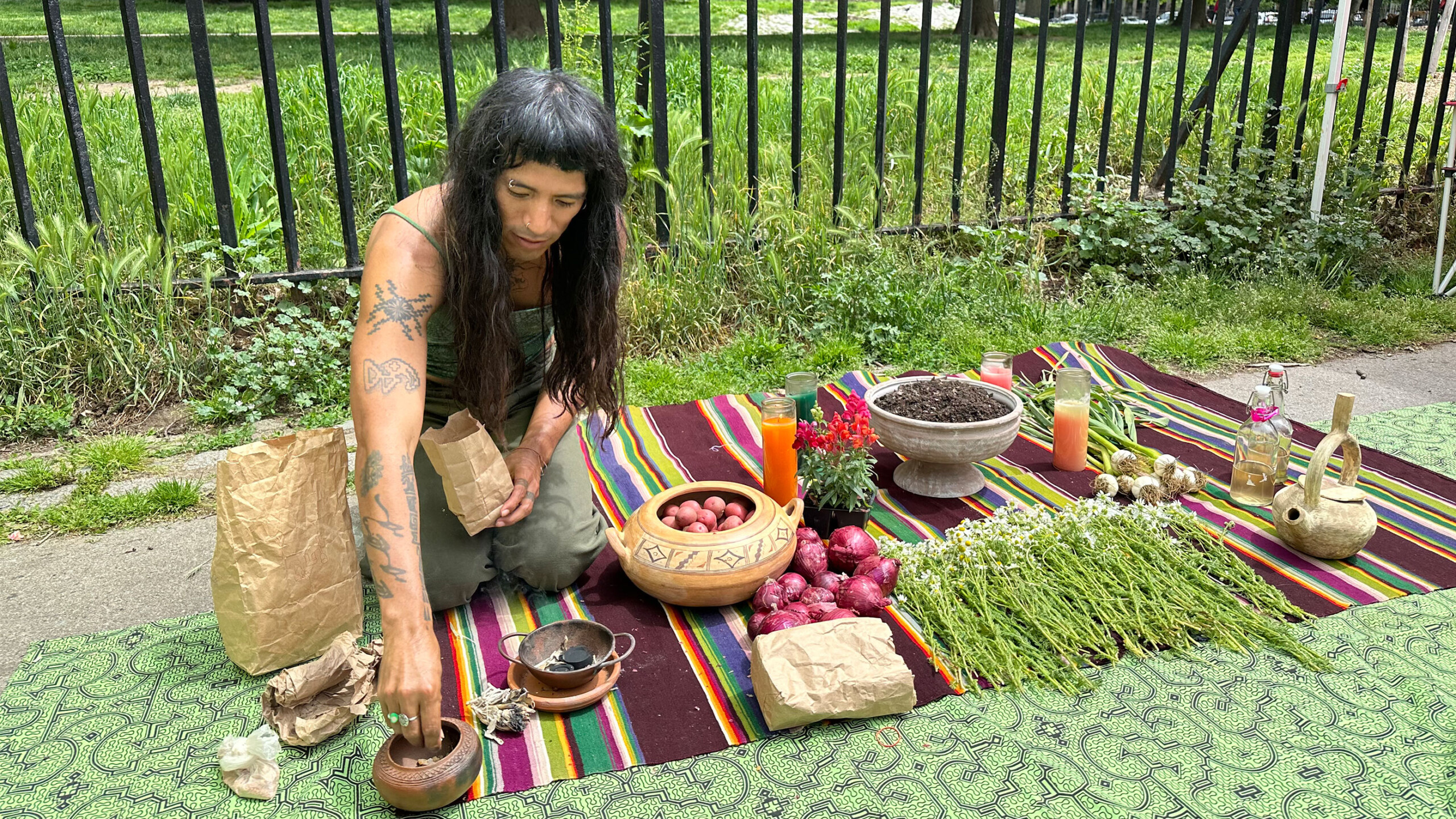 A person with long, dark hair and visible tattoos is kneeling on a colorful blanket outdoors, surrounded by a variety of items, possibly for a ritual or ceremony....