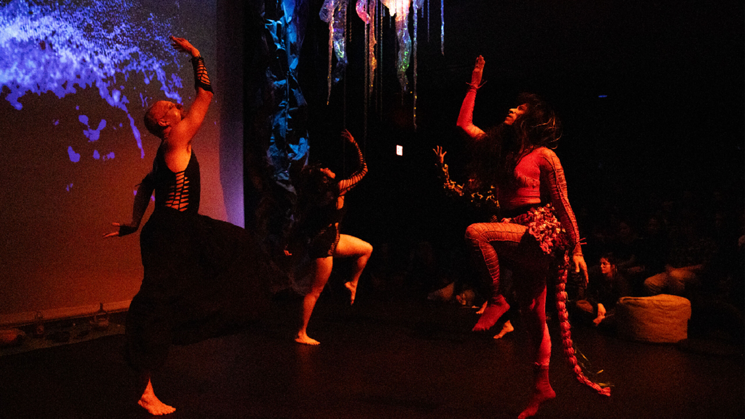 A group of performers dressed in elaborate costumes engage in a dynamic dance routine on stage. The lighting is dim, creating an intimate atmosphere, while a projection...