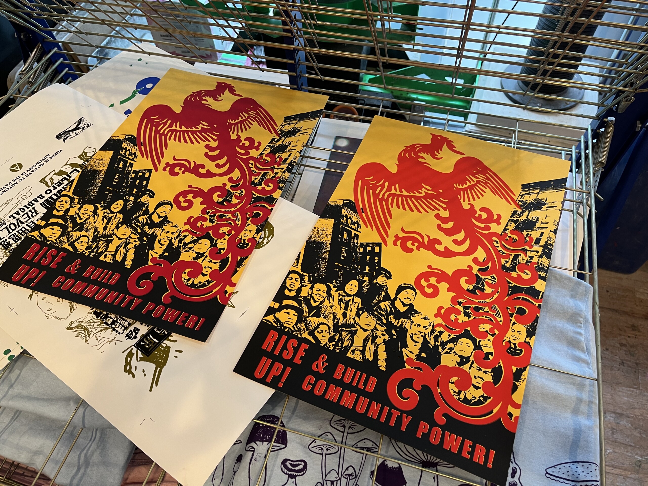 Photograph of two identical posters drying on metal racks, depicting a red phoenix flying above a black and yellow print of people standing amid buildings. The...