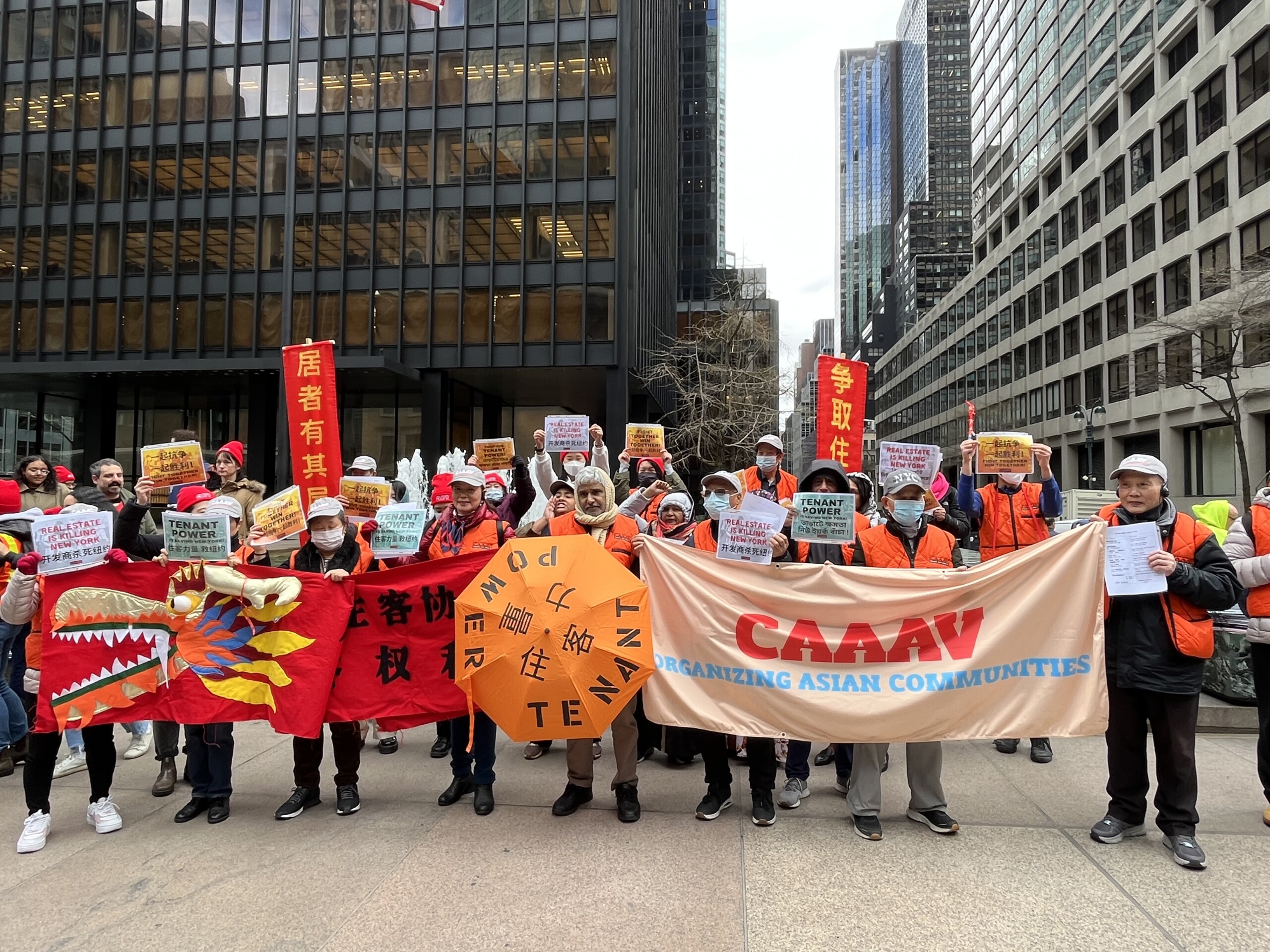 Photograph of Asian elders and allies standing outside amid tall buildings in NYC, wearing winter clothes and face masks. They are holding banners and signs written...