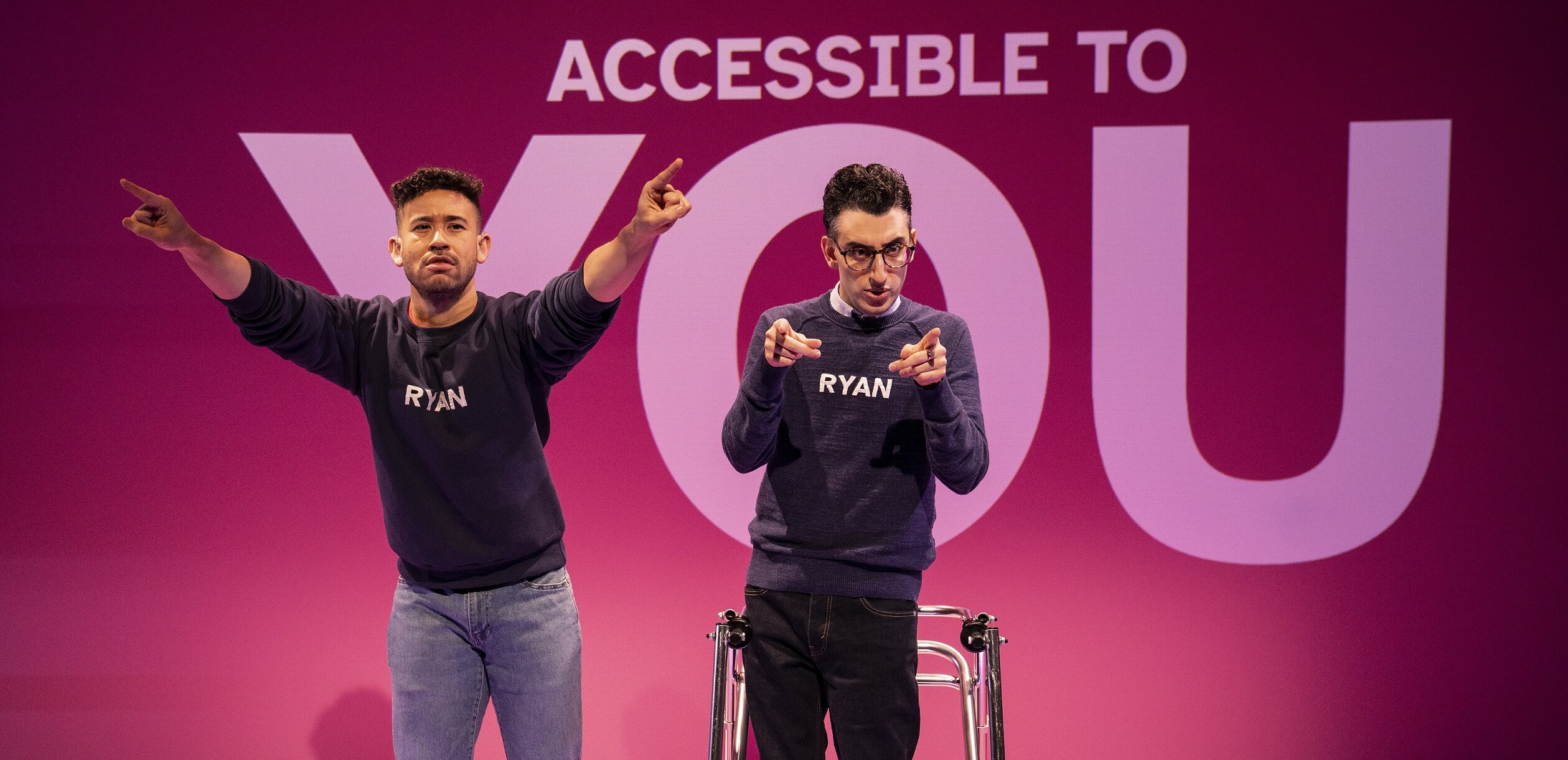 Photo of two men, one using a walking support device, on stage. Behind them, text on the wall reads Accessible to YOU