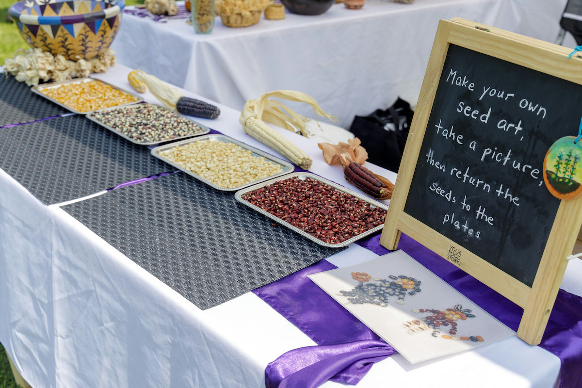 A table covered with a white tablecloth and decorated with a purple silky runner is set up for a seed art activity. Several trays filled with different types of...