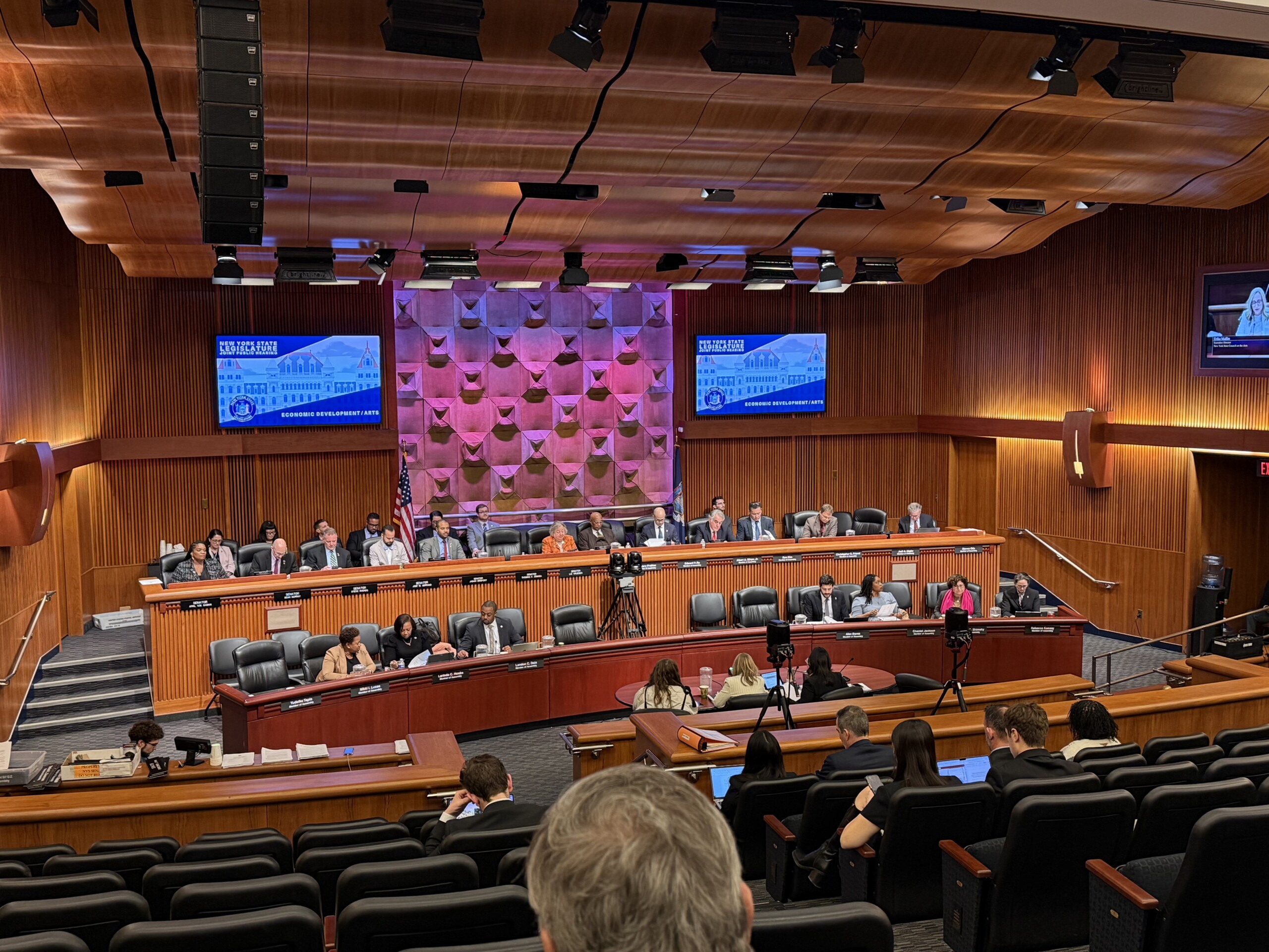 Photograph of government representatives of varying genders and races seated in a large town hall, facing a round table and audience seats. Behind the representatives...