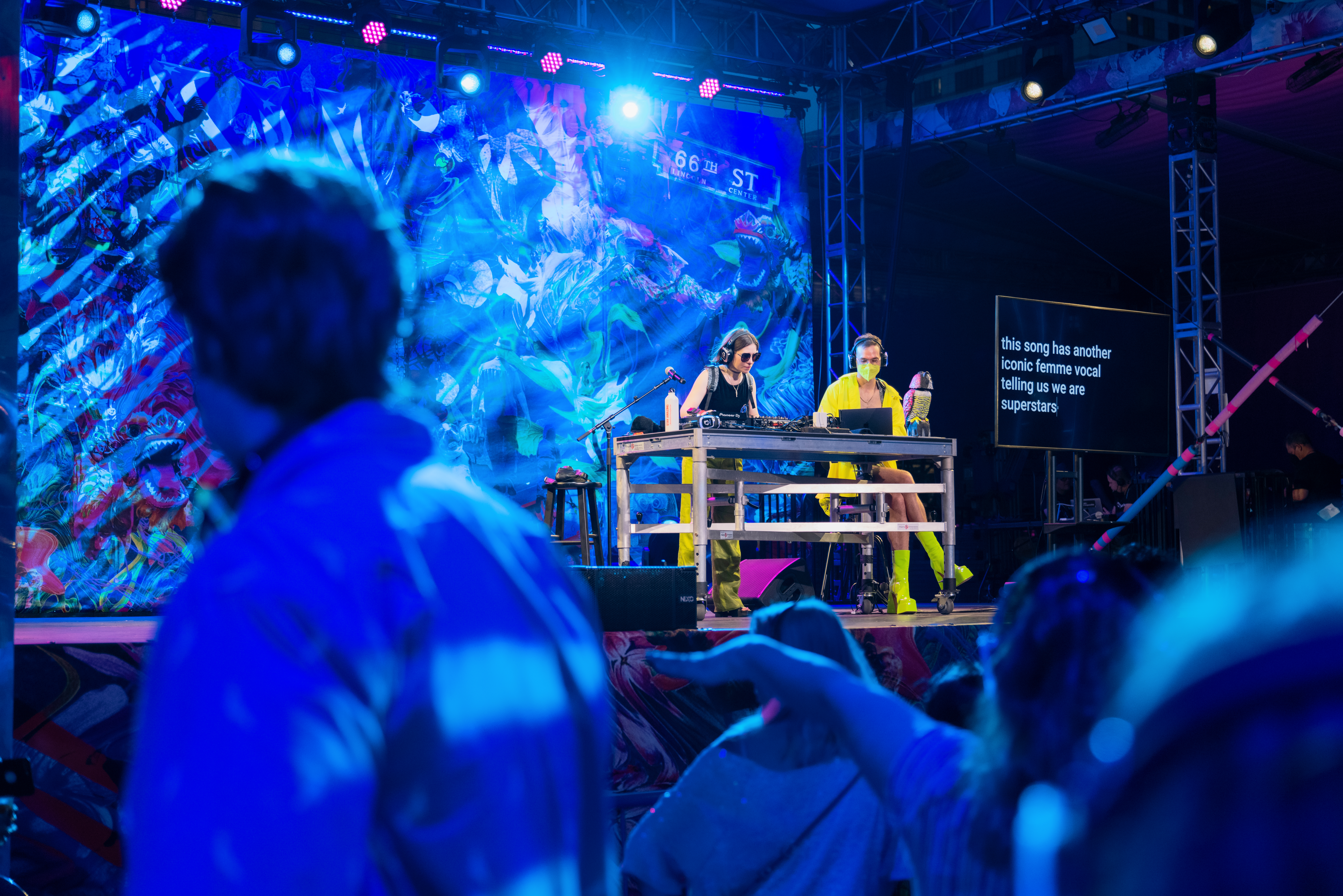 On a large stage, a white femme DJ is at work organizing the sounds being sent to silent disco headphones. Next to them, a scruffy white artist wearing chunky neon...