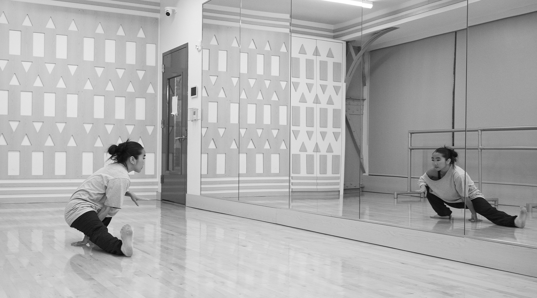 Black and white photograph of a dancer with her hair in a bun stretching in a squat with one extended leg. Her reflection is in a large mirror running along the...