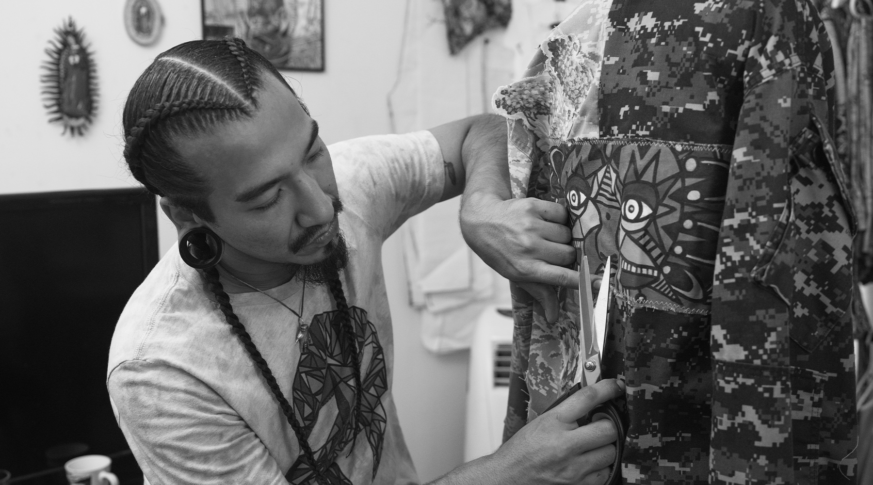 Black and white photograph of a fashion designer wearing ear gauges and French braids using scissors on a patterned garment inside a room with art on the walls.