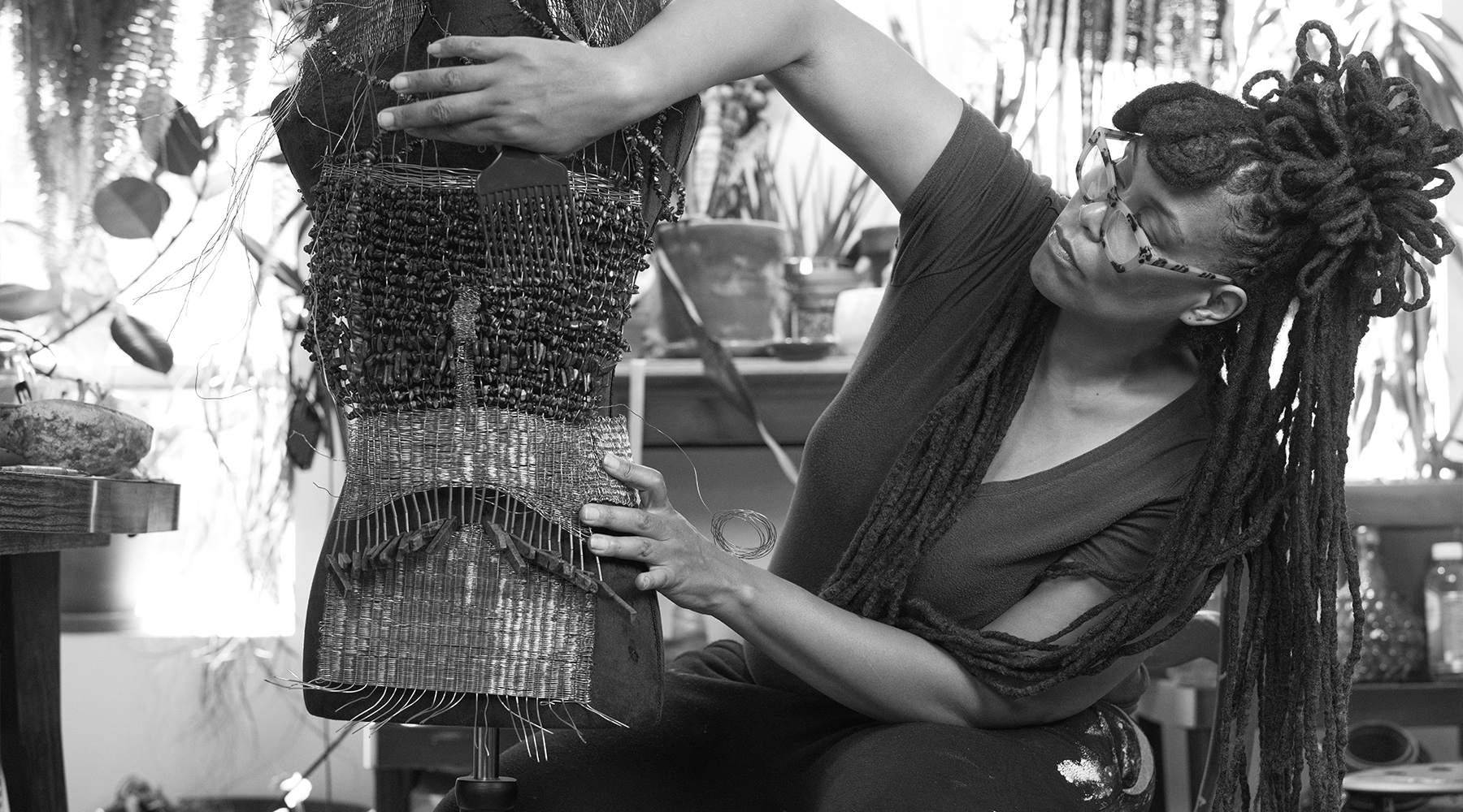Black and white photograph of an artist wearing glasses creating a sculptural textile piece on a mannequin inside a room filled with plants.