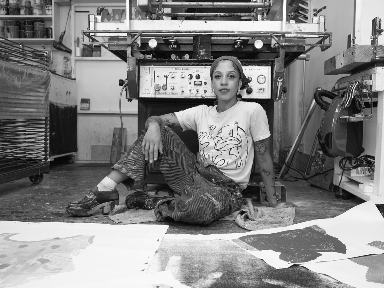 Black and white photograph of an artist wearing a hair bandana and printed t-shirt posing with her elbow resting on her knee. Her pants and shoes are splattered...