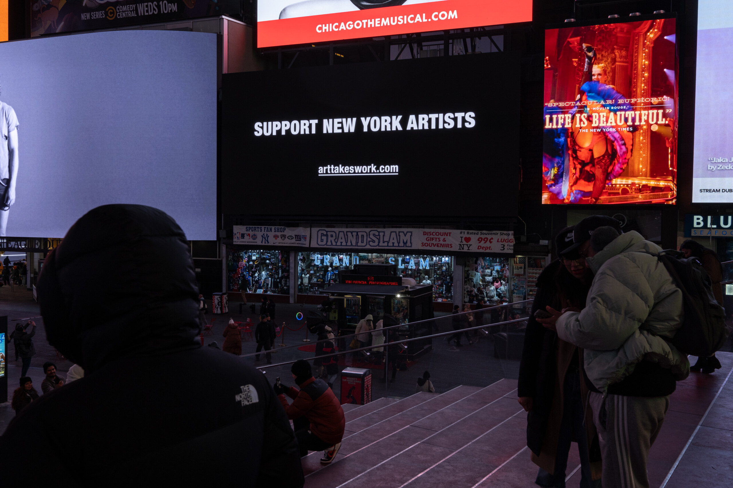 A photo of a screen in Times Square. It is all black with white text that says, Support New York Artists. ArtTakesWork.com.