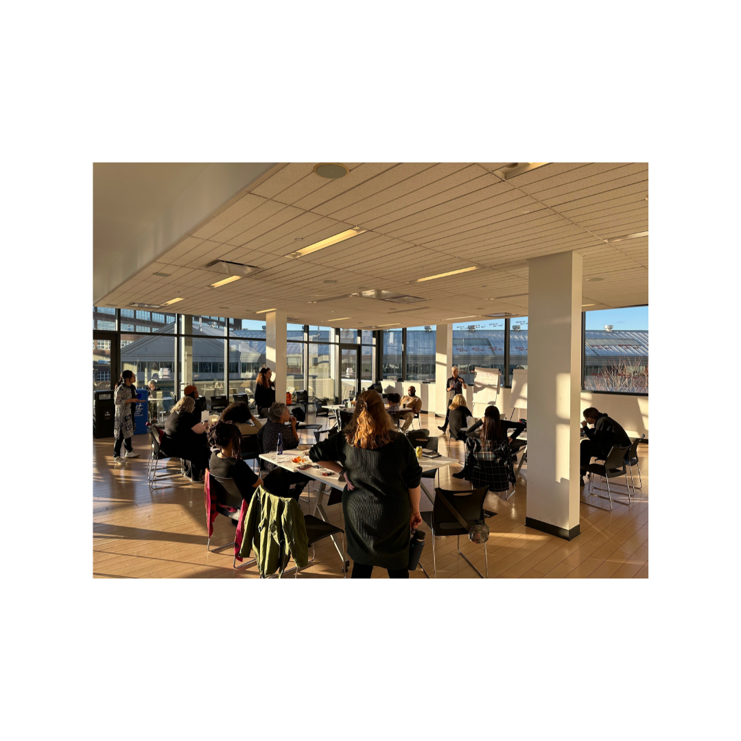 Photograph of roughly 16 people of varying races and genders sitting at tables set up in a semicircle. The tables are covered in black tablecloths, papers, office...
