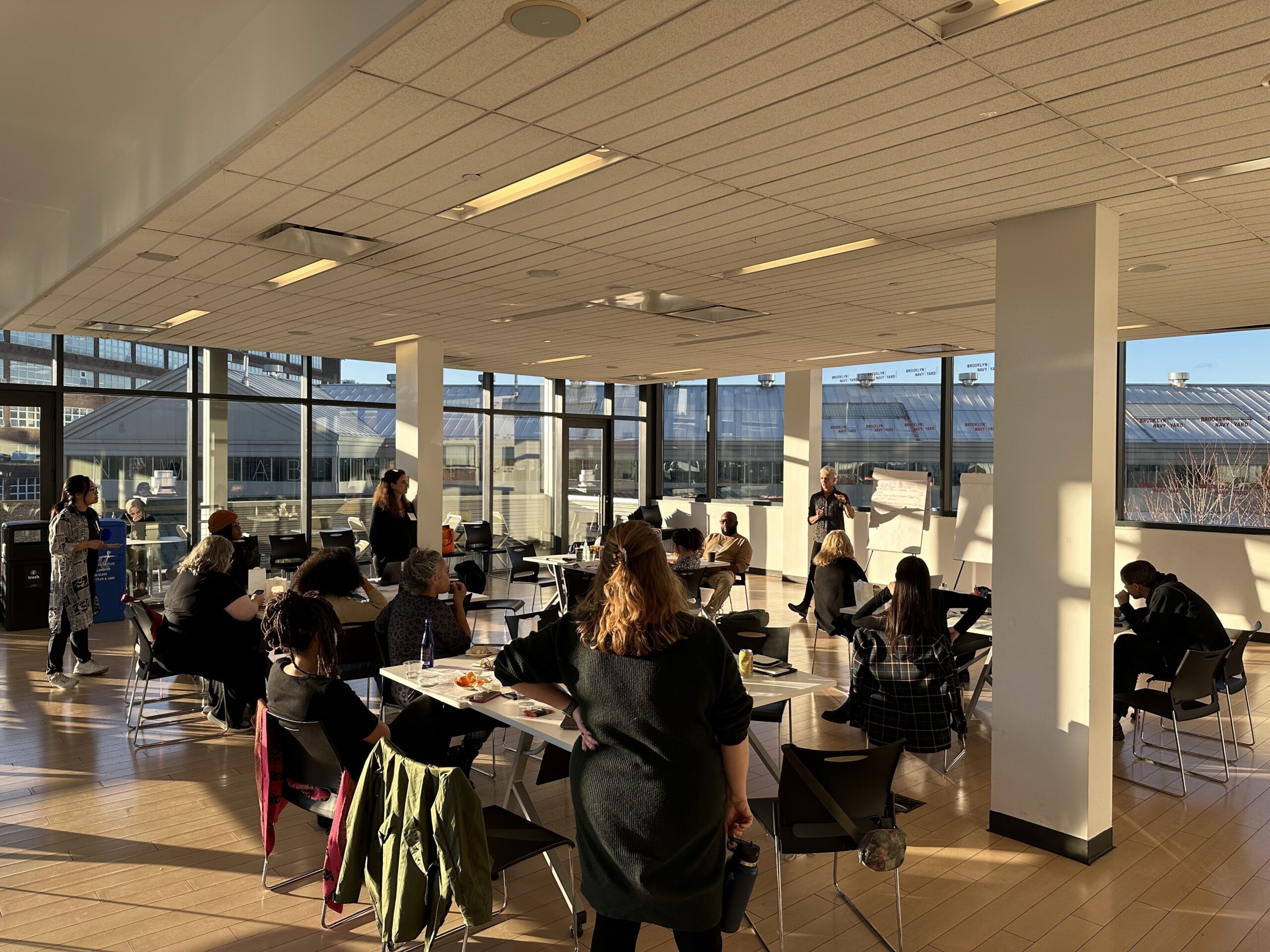 Photograph of a group of people casually spread out in a room in seated or standing positions. Most people have their back towards the camera as they face a standing...