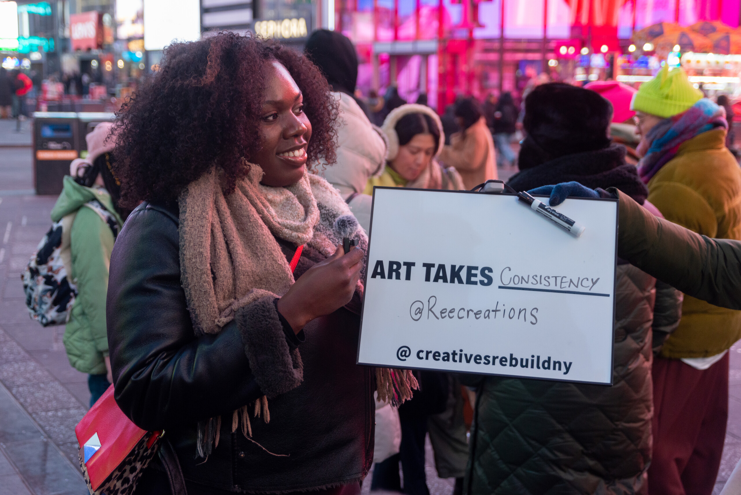 A photo of a Black woman with curly shoulder-length hair is holding a small micrphone and a sign outside in Times Square. The sign says, Art Takes blank. This artist...
