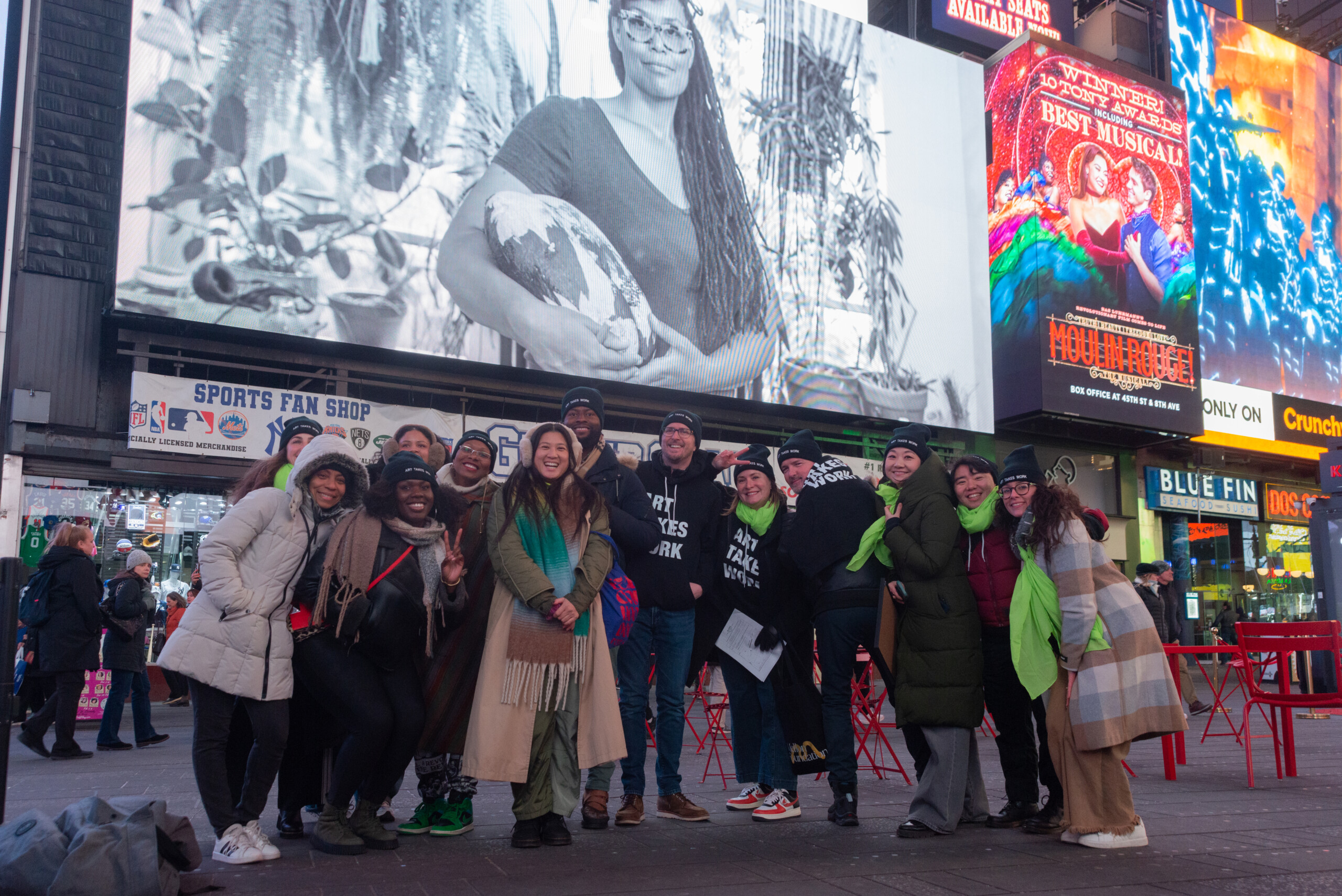 A group of thirteen people posing on an urban street in front of a large screen with the phrase Art Takes Work written in bold white type on a black background....