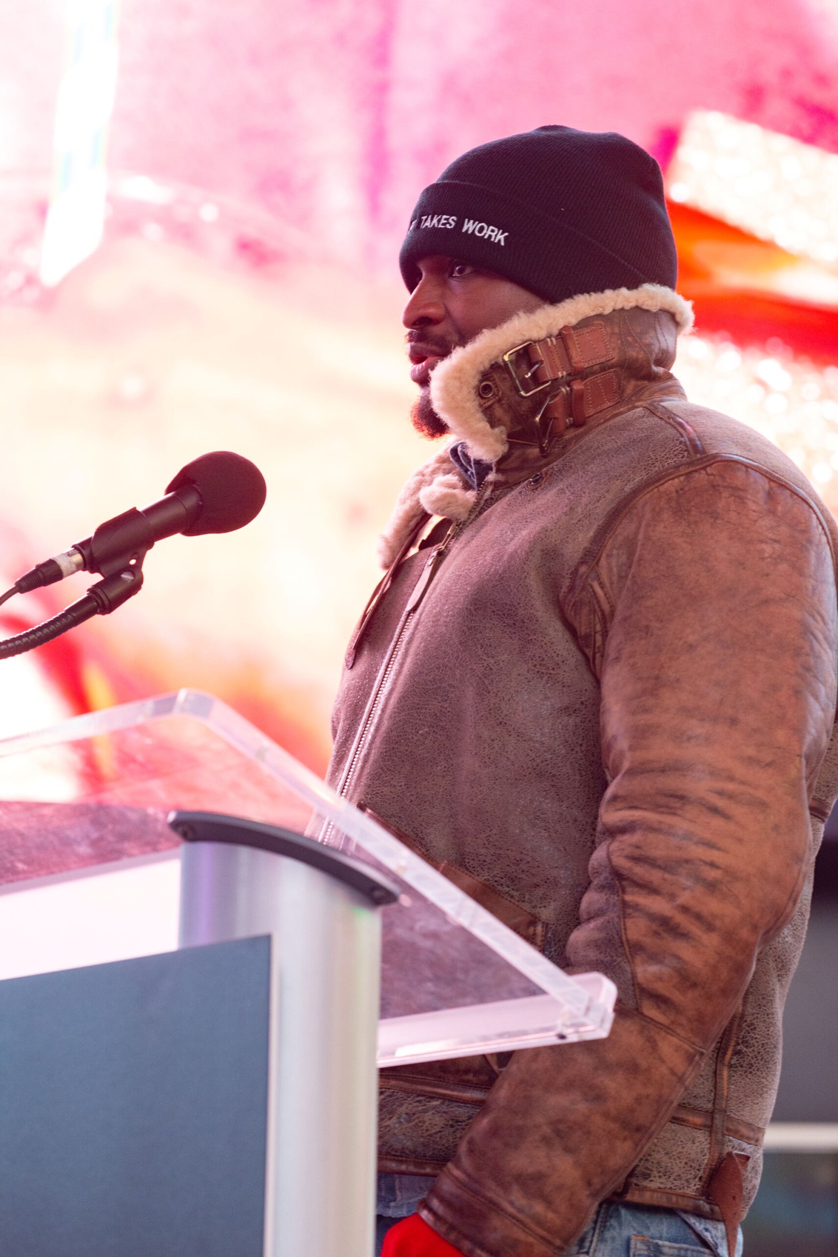 CRNY program artist Russell Craig speaks into a microphone at a podium. He is a black man wearing a brown leather jacket and a black beanie that says ART TAKES...