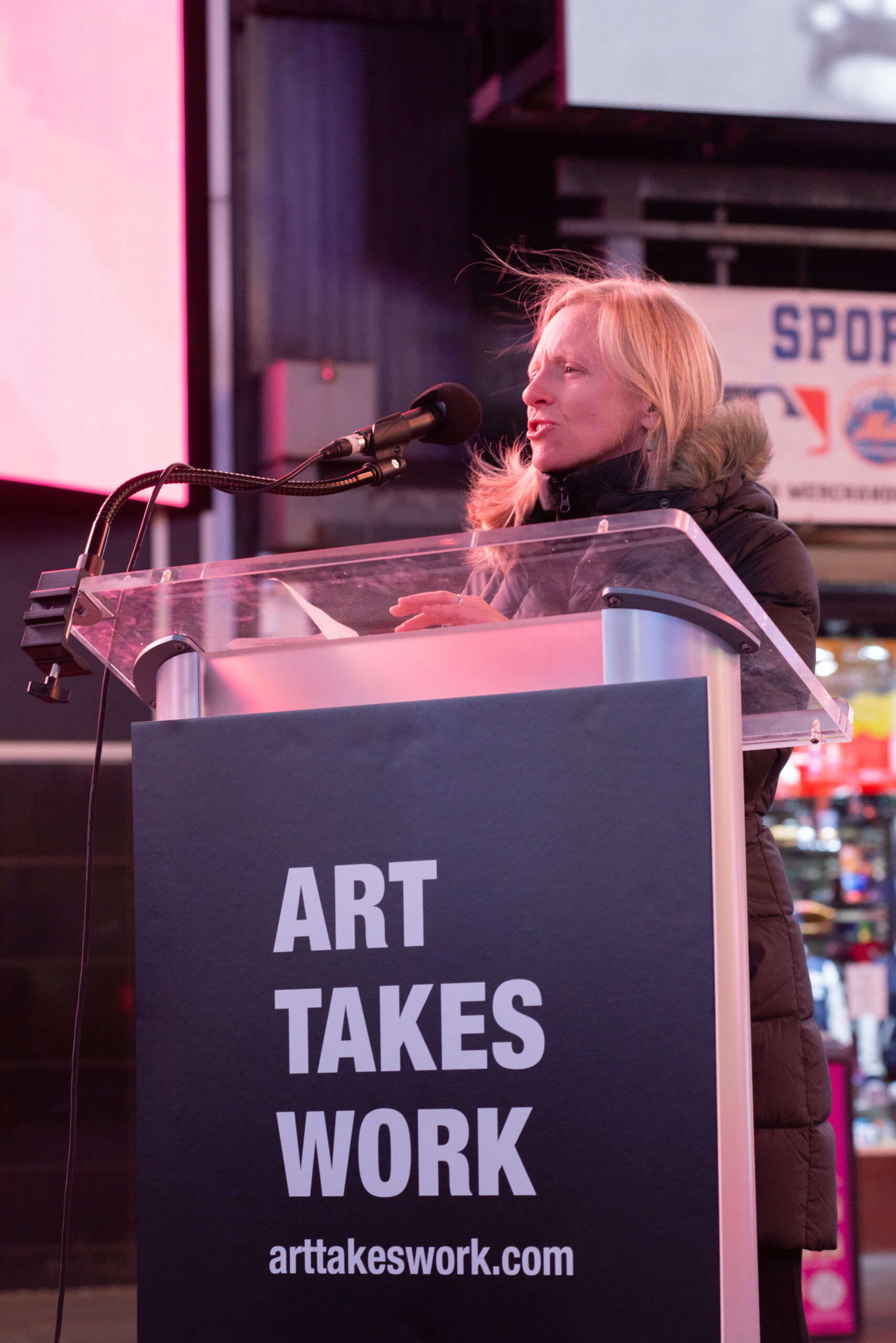 A photo of a white woman speaking into a microphone at a podium. Wisps of her blond hair are blowing in the wind in Times Square.