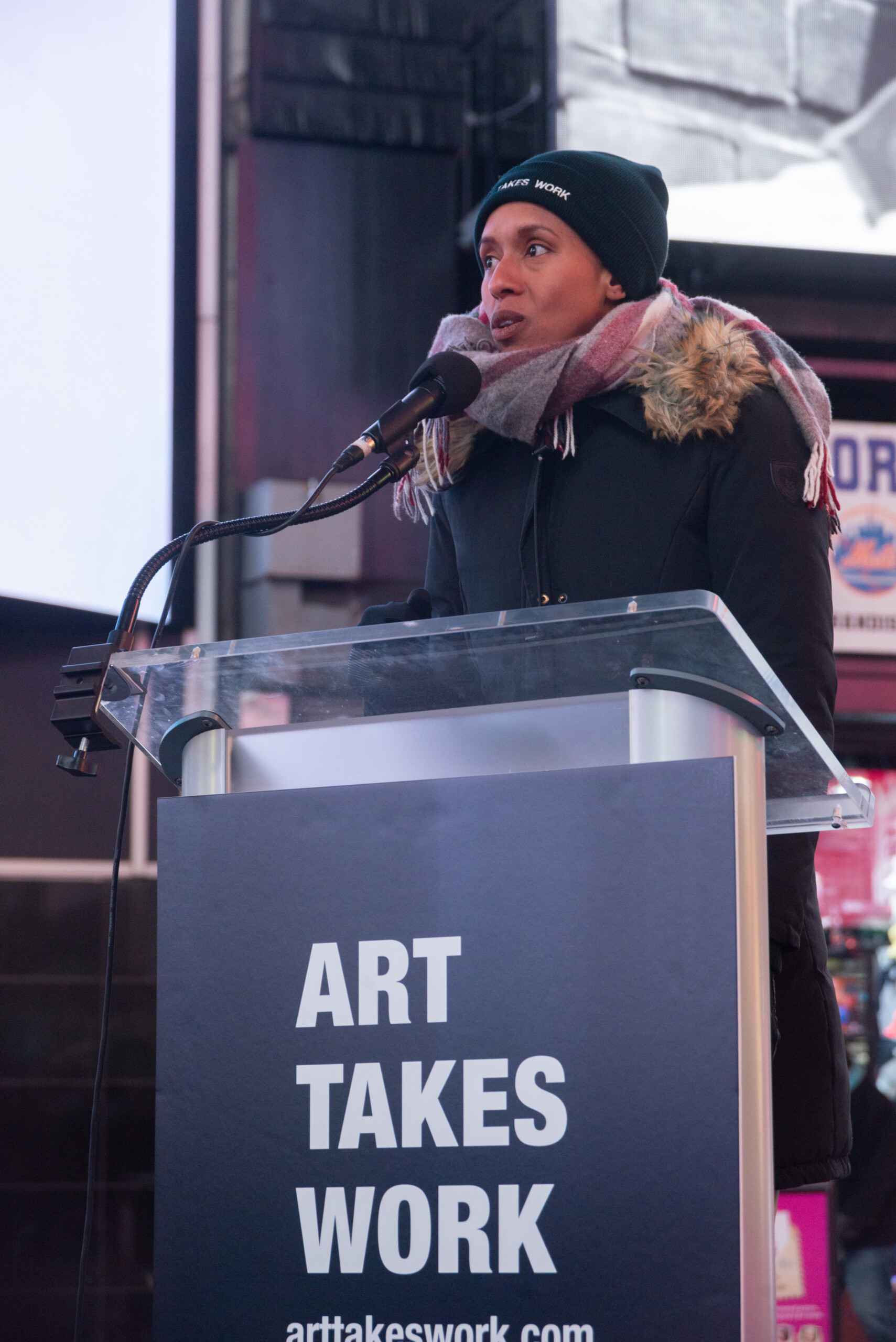 A photo of a Black woman speaking into a microphone at a podium. She wears a heavy winter coat, a scarf, and a beanie hat that says, Art Takes Work.