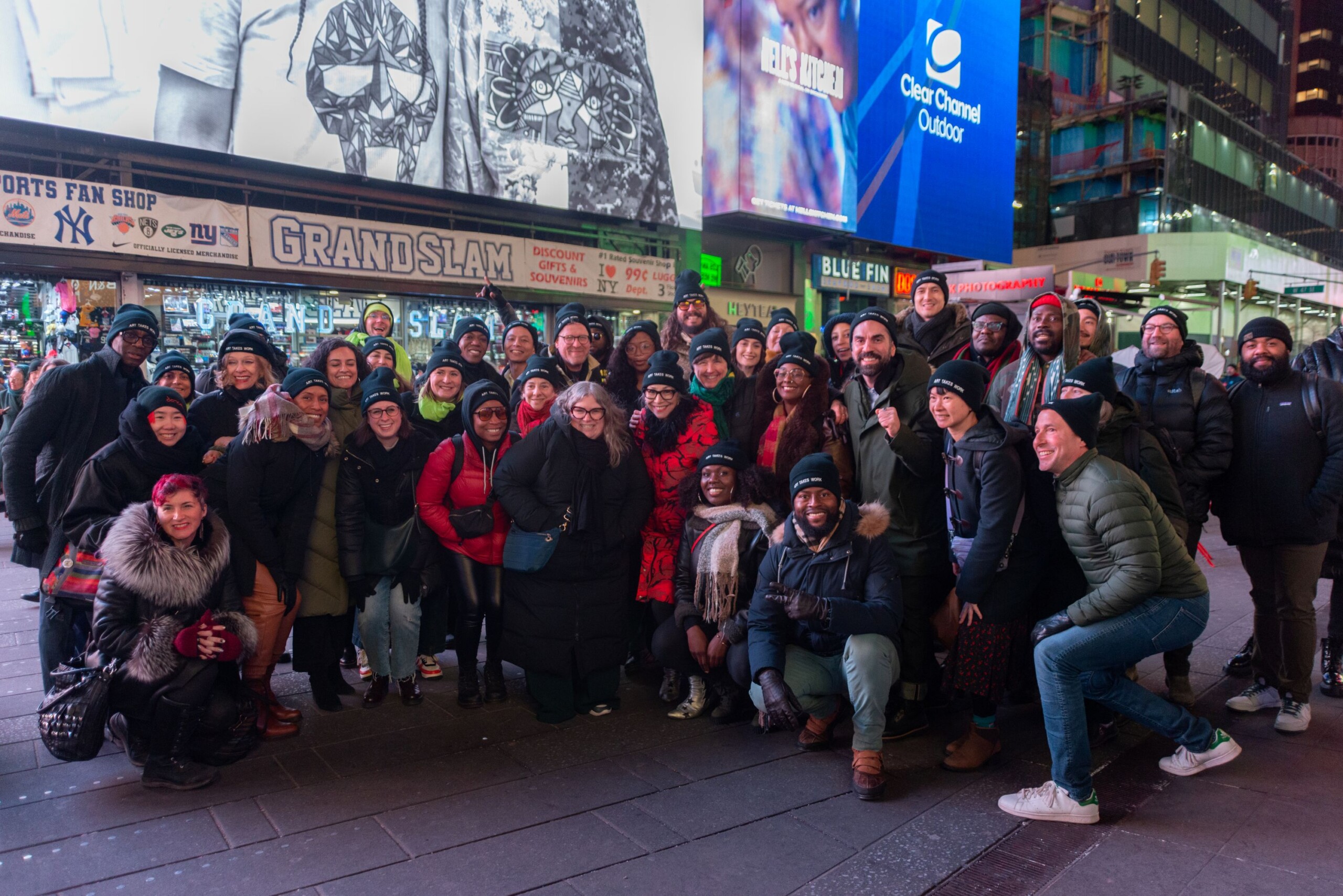 A large group of people smiling and posing in an urban plaza at night. They are dressed warmly, and many are wearing a black hat with the phrase Art Takes Work...