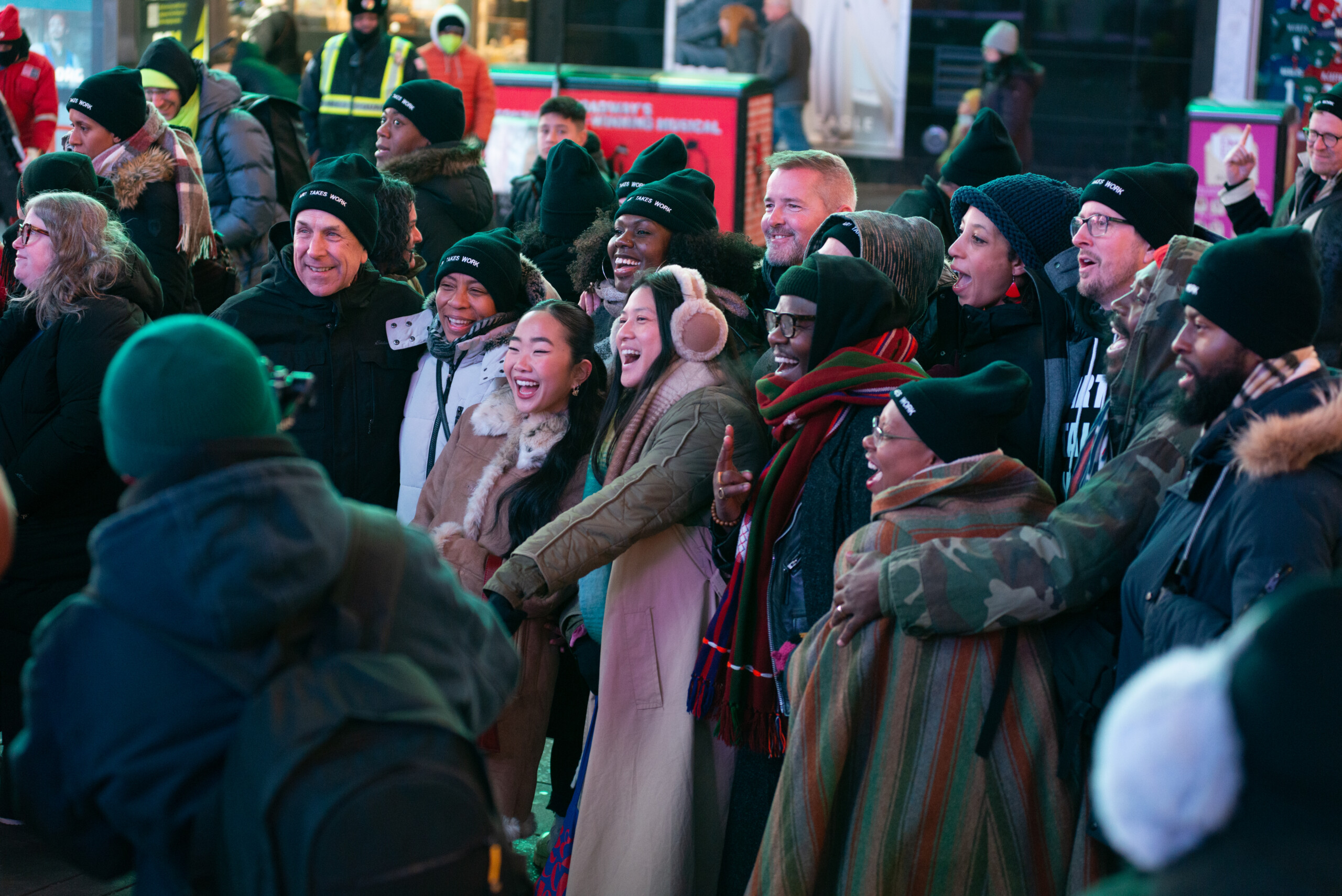 A group of artists are huddled together for a group photo. They are outside in Times Square. Many wear black beanie hats that say, Art Takes Work.