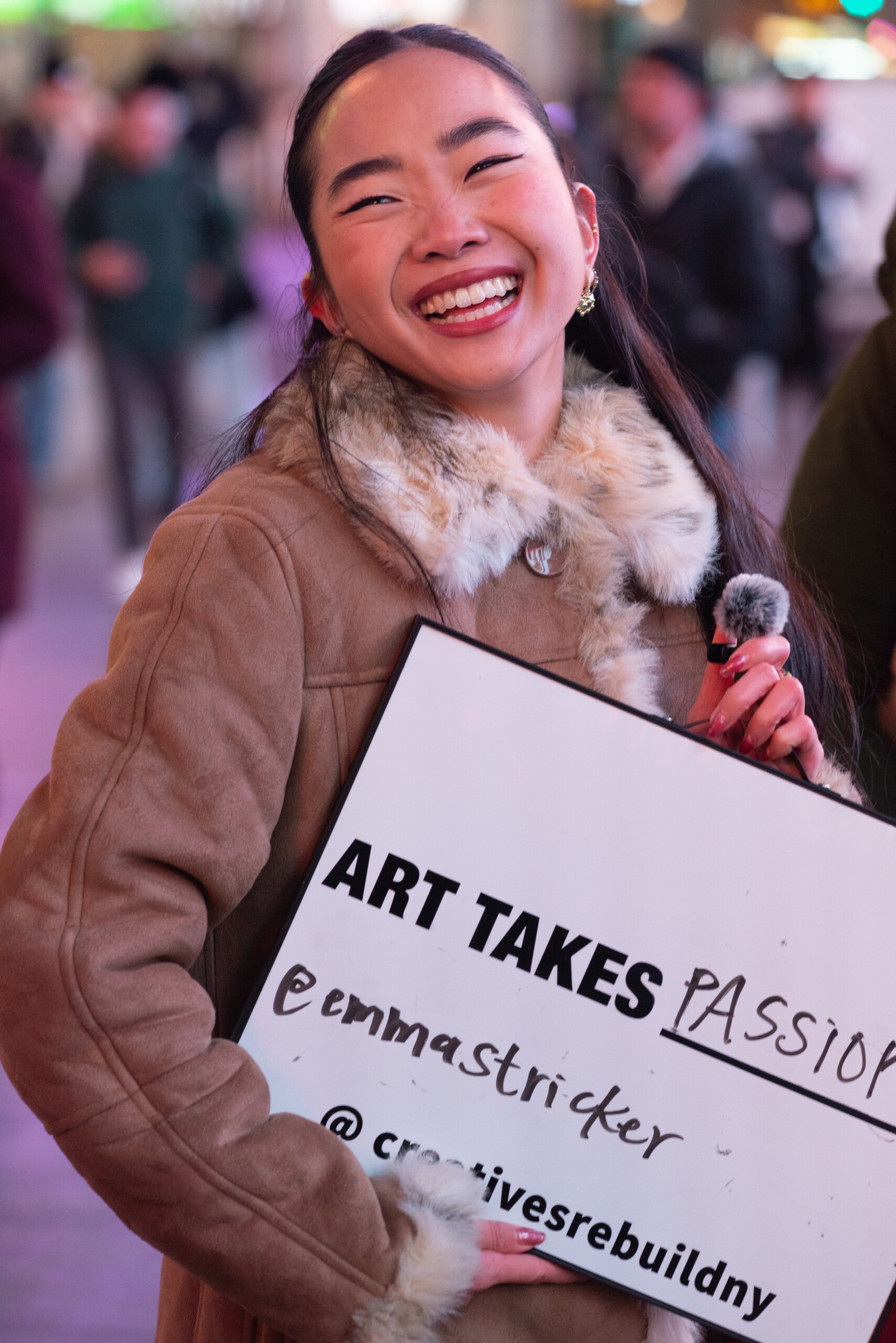 A photo of an Asian American woman smiling widely and holding a small microphone and sign. The sign says, Art Takes blank. This artist has filled it in to say,...