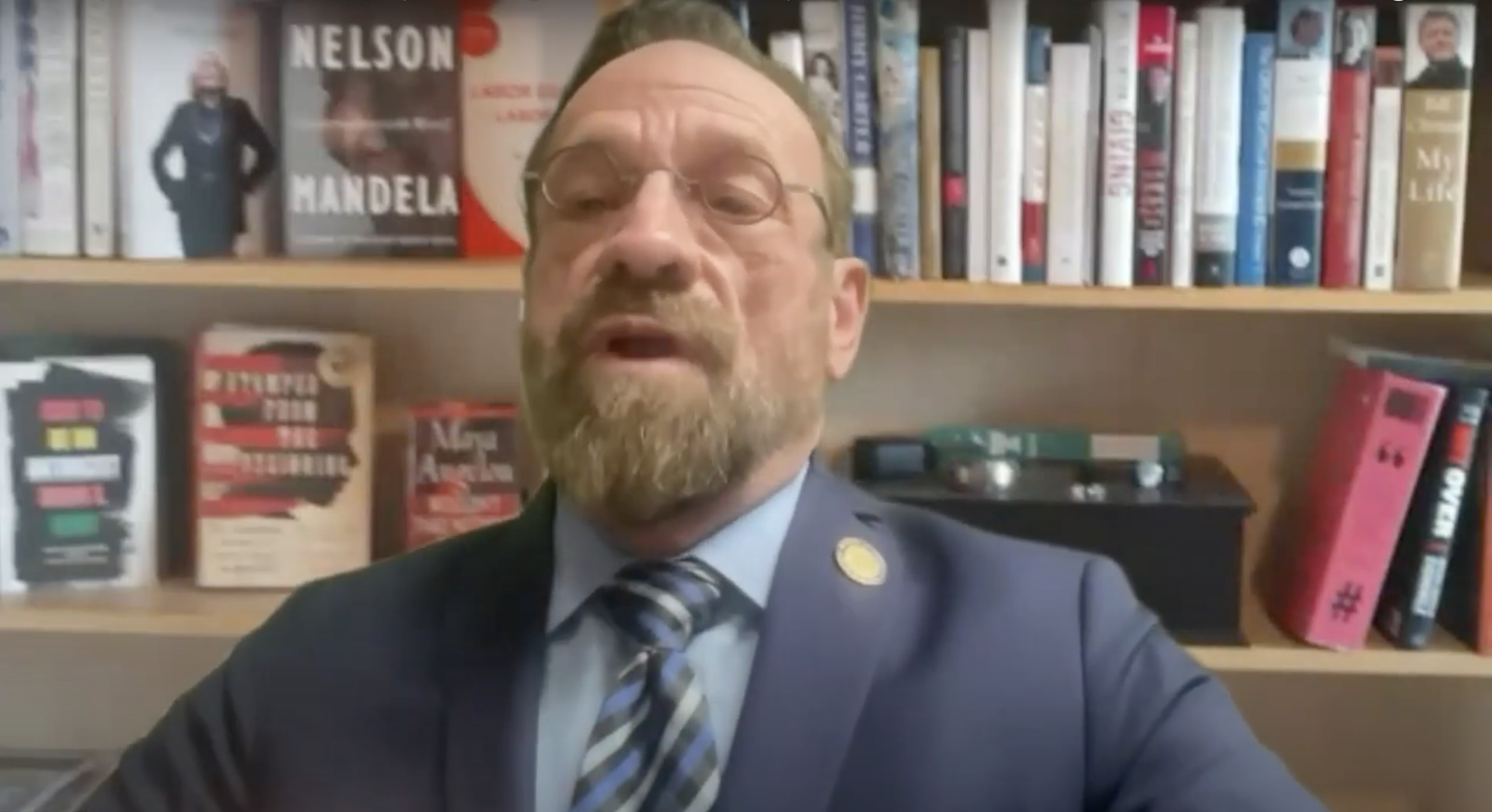 A white man with glasses, a beard, and a bald head wearing a dark blue suit, striped tie, and a lapel pin is speaking while seated in front of a bookshelf. The...