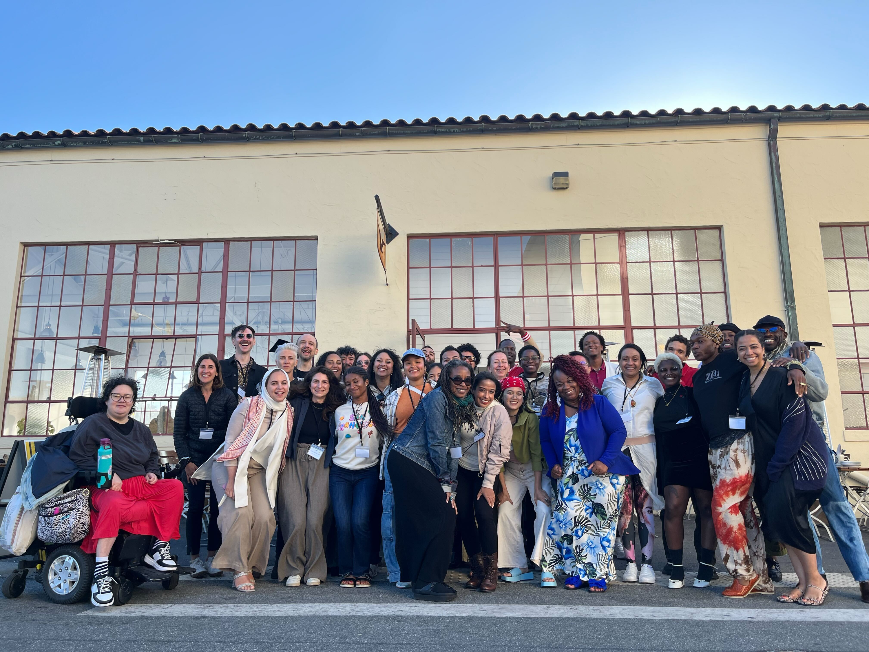 A large group of diverse people, including individuals of different ages, races, and abilities, pose together outside a beige building with large windows. Everyone...