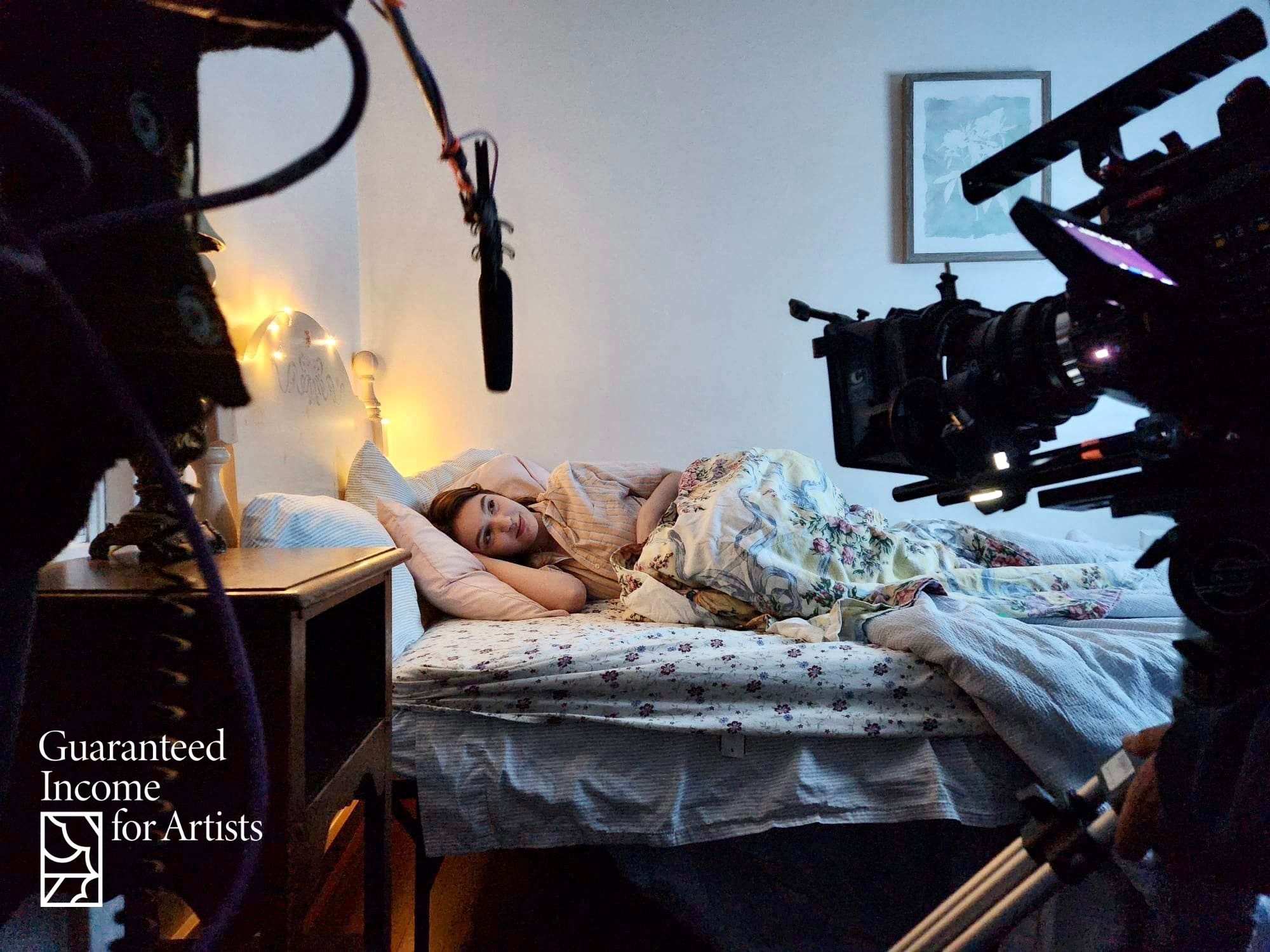 Behind-the-scenes photograph of a film set where an actor is lying awake in a bed. The bed has floral sheets and blankets and is inside a room with white walls,...