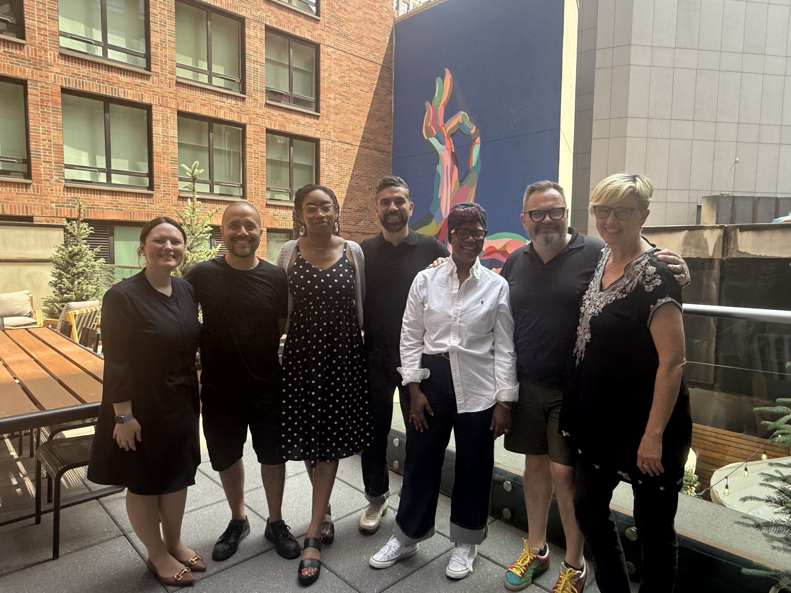 Seven smiling people stand on a balcony facing the camera. Behind them is a multicolored mural and several glass windows on the side of a brick building.